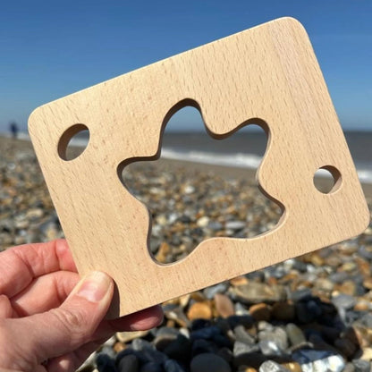 Wooden starfish-shaped toy held by a hand on a pebble beach with blue sky.