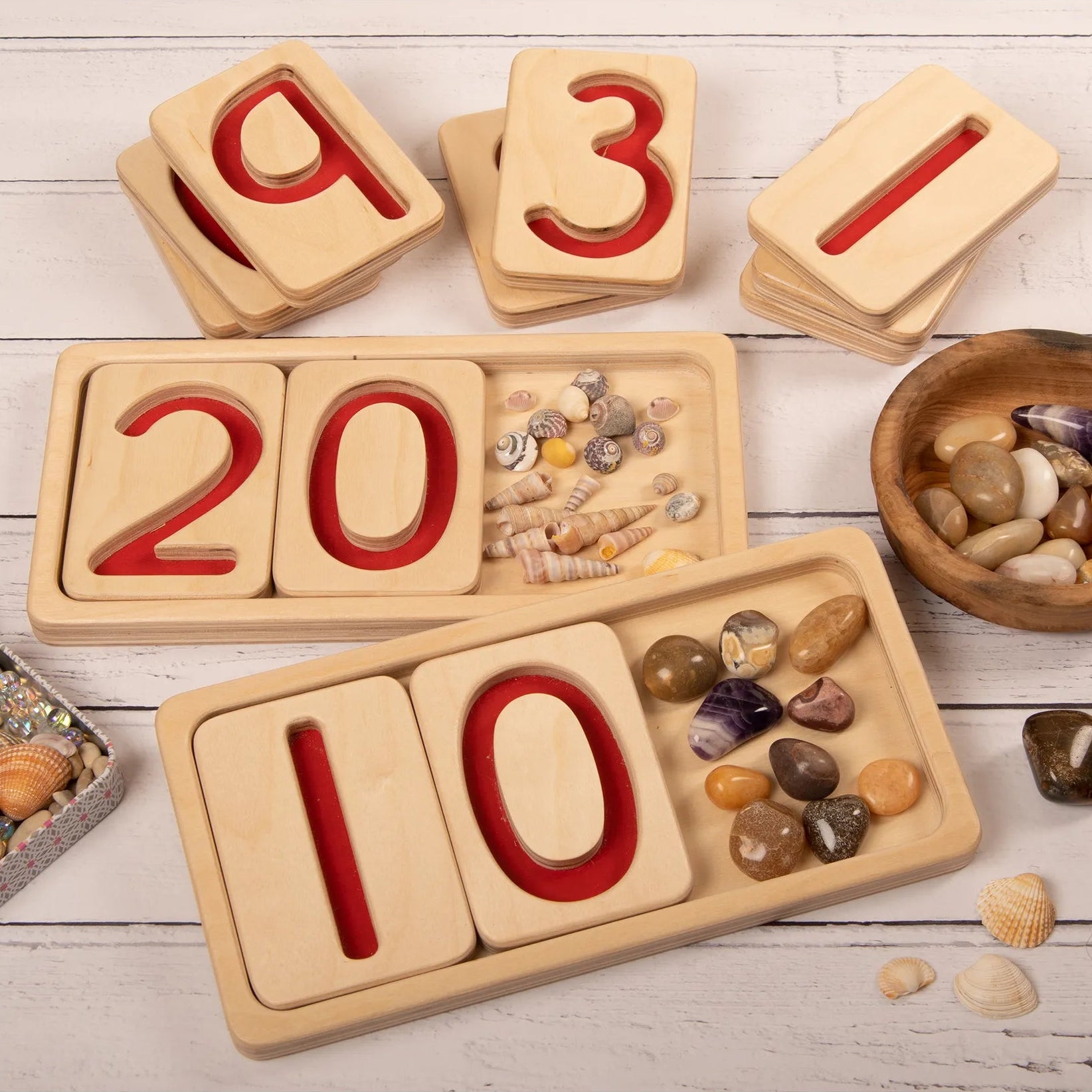 Wooden number trays with red numeral indents on a wooden surface with loose parts to count from 10-20 and 0-10.