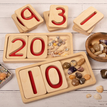 Wooden number trays with red numeral indents on a wooden surface with loose parts to count from 10-20 and 0-10.