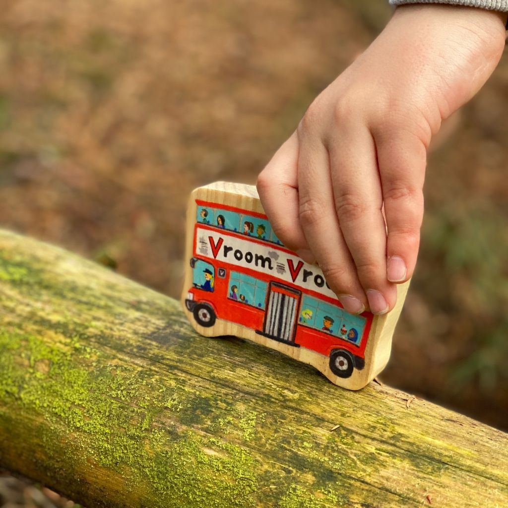 Hand holding a wooden toy bus with 'Vroom Vroom' text on a natural background
