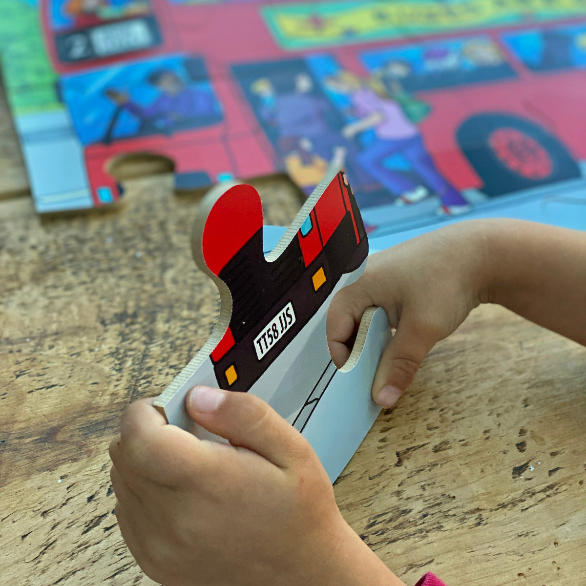 Child playing with a jigsaw puzzle piece on a wooden table with a colorful bus in the background.