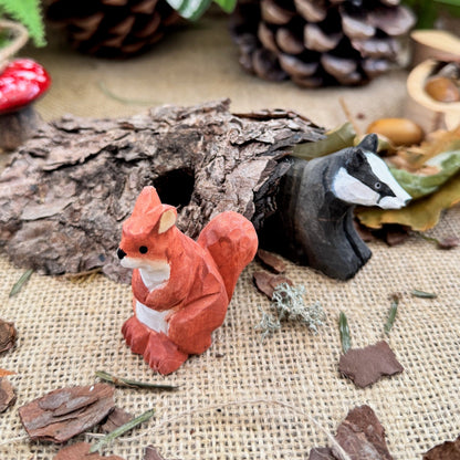 Small red squirrel figurine with a white label on a rustic background with pinecones and leaves.