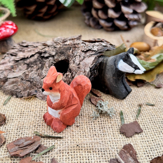 Small red squirrel figurine with a white label on a rustic background with pinecones and leaves.