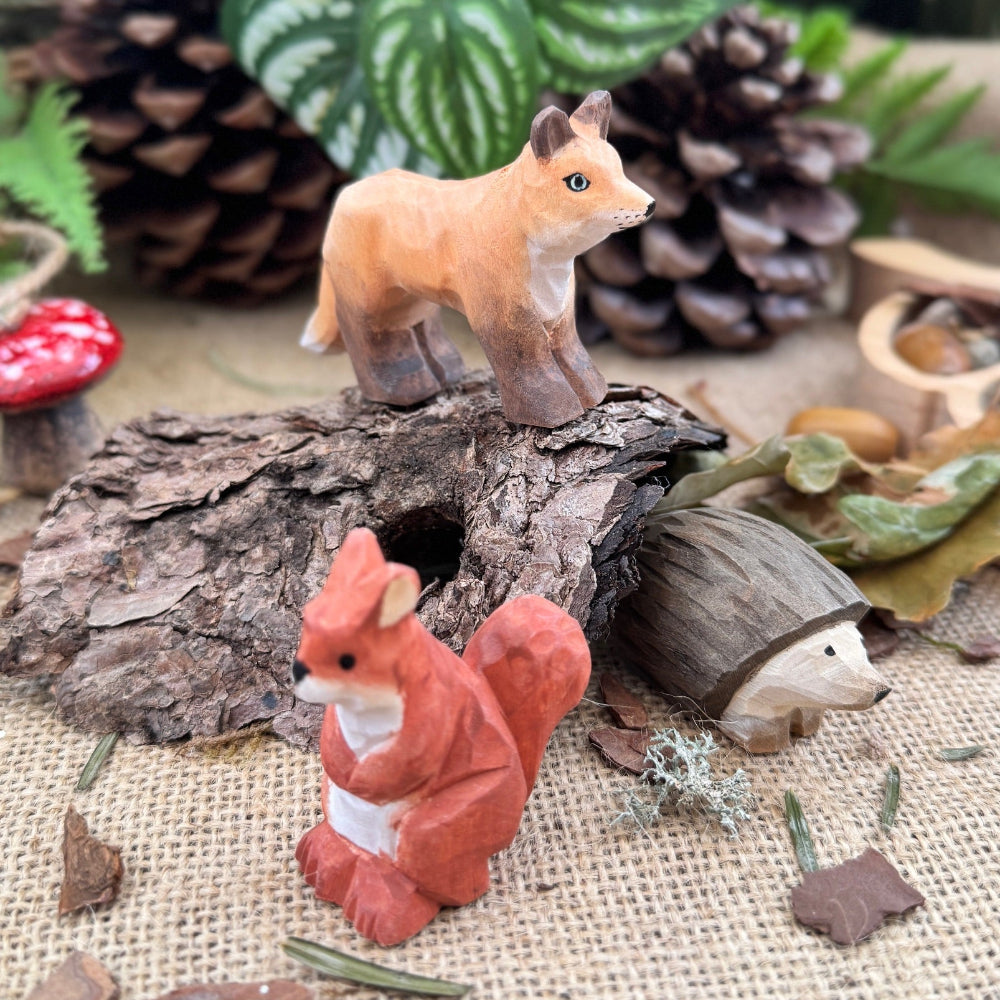 Miniature fox, hedgehog and squirrel figurines on a wooden log with pine cones and plants in the background.