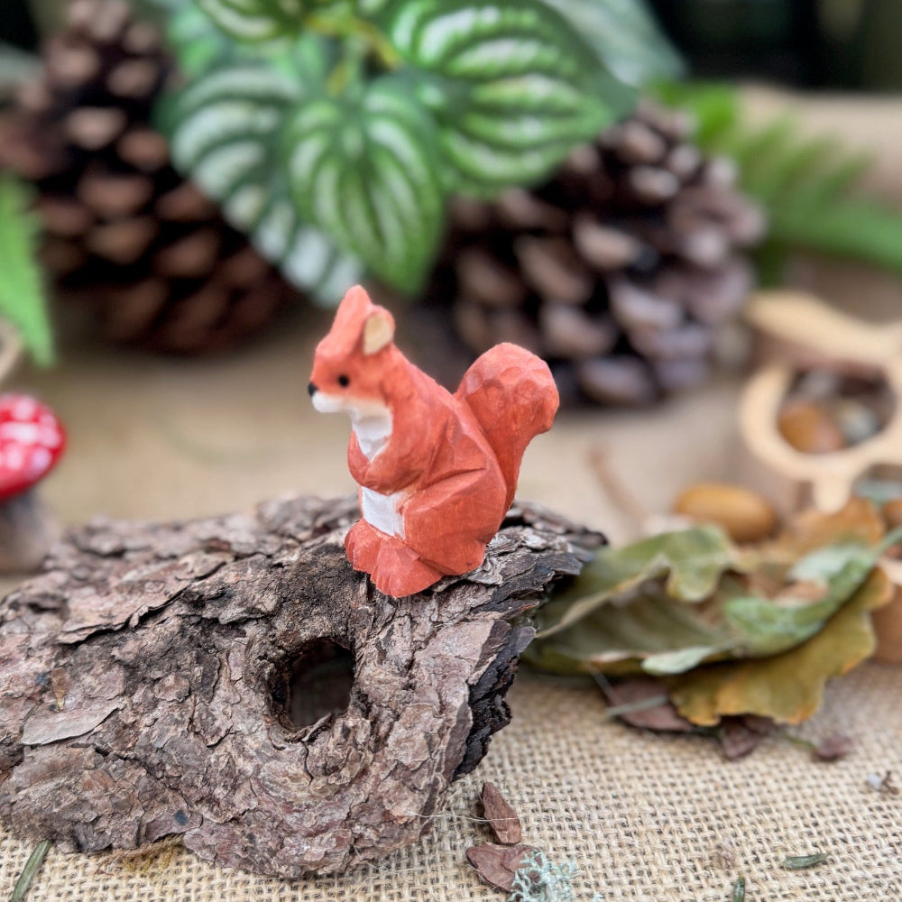 Small orange squirrel figurine on a piece of wood with pinecones and leaves in the background.